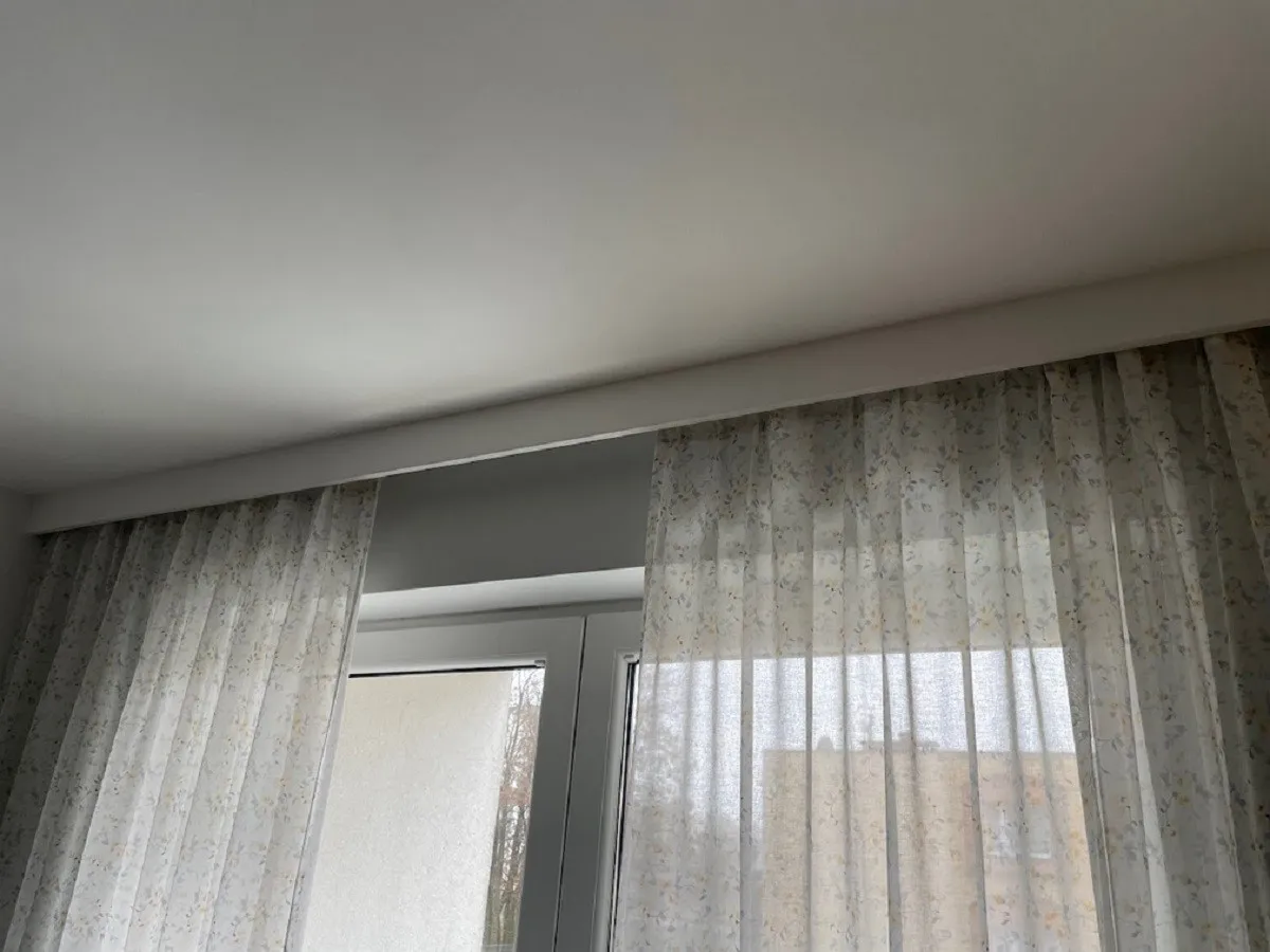 Jasne, oto propozycja:
Jasny, kwiecisty materiał zasłania okno, a nad nim widać gruby pasek pod sufitem.