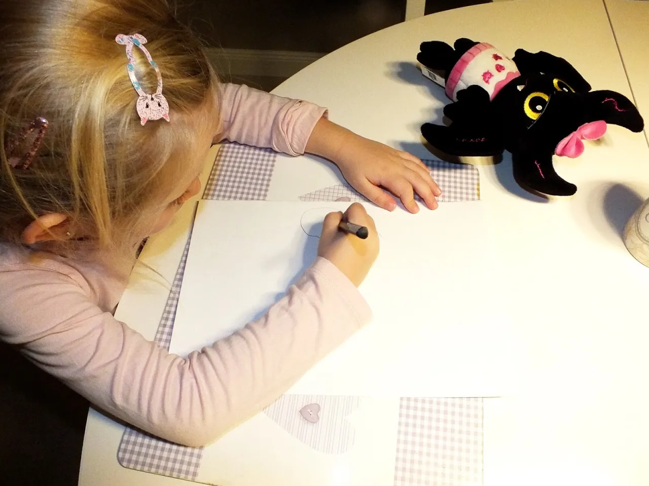 Dziewczynka rysuje, obok leży czarny pluszowy kotek. Jak opisać pluszowego misia?