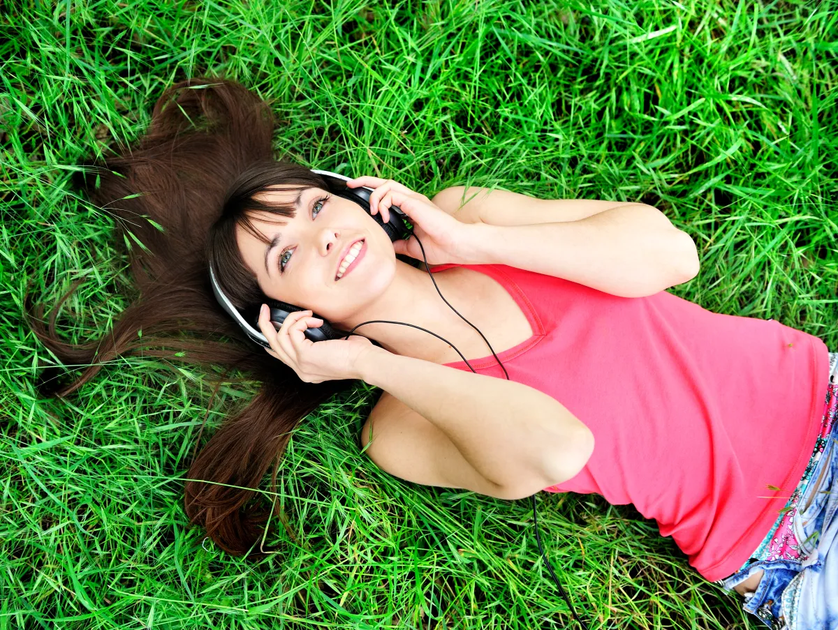 Uśmiechnięta dziewczyna w słuchawkach leży na trawie. Zastanawia się, gdzie może wysłać swoje teksty piosenek.