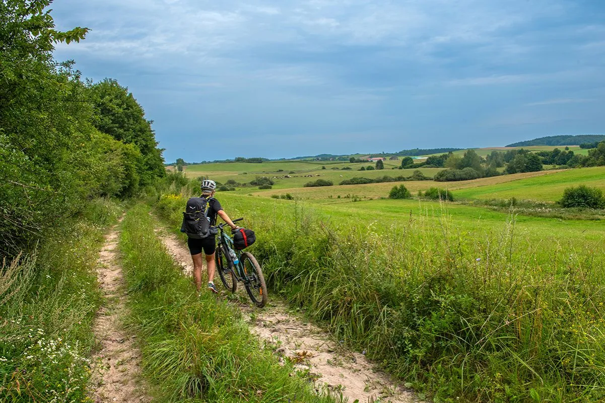 Kobieta z rowerem na piaszczystej trasie rowerowej wokół jeziora, otoczona zielonymi łąkami i drzewami.