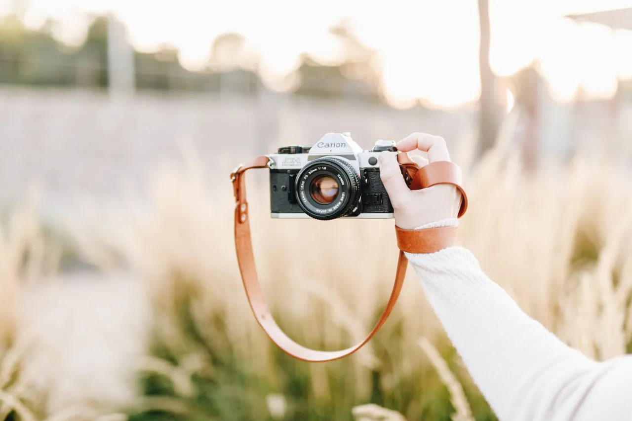 Dłoń trzyma aparat Canon AE-1 z brązowym paskiem. Chcesz wiedzieć, jak zrobić ładne zdjęcia? Zacznij od dobrego sprzętu!