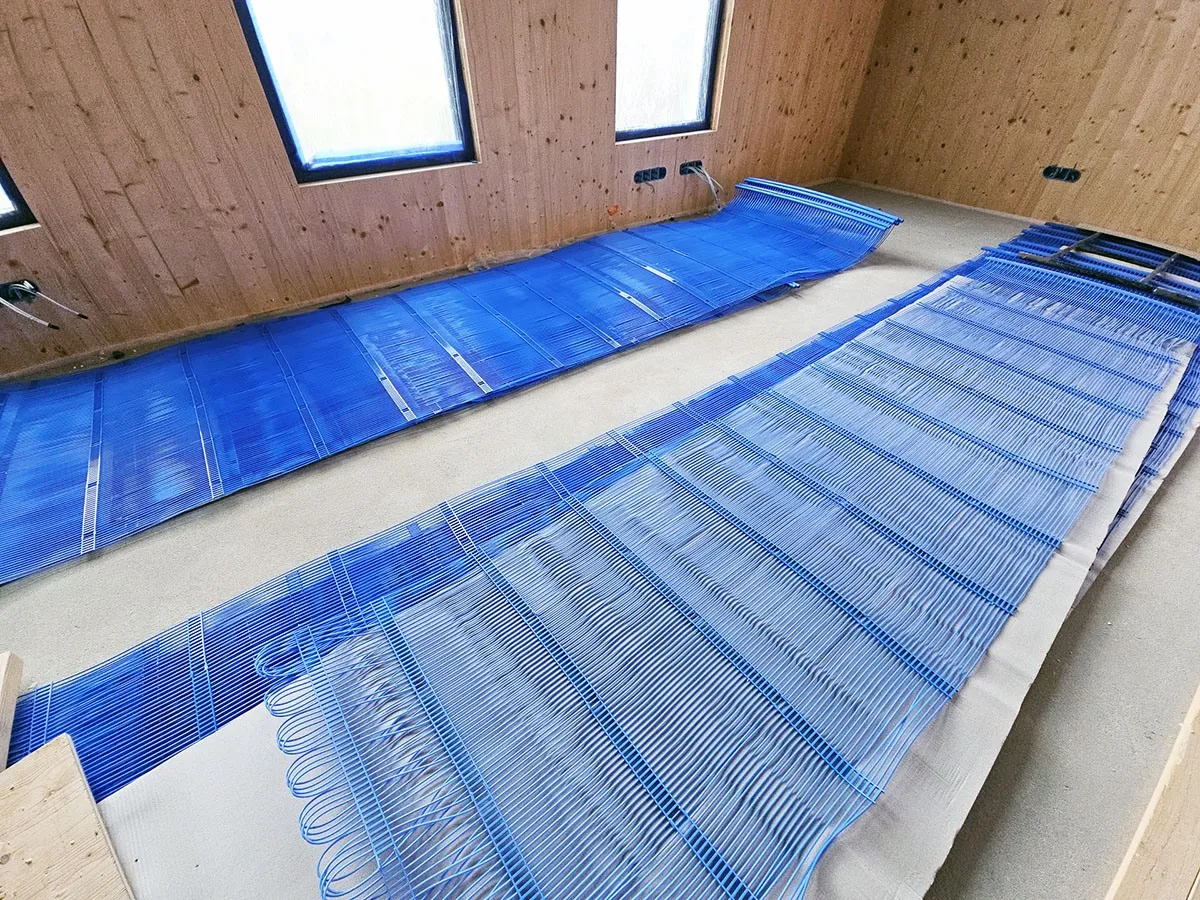 Dwa niebieskie maty kapilarne rozłożone na podłodze w drewnianym pomieszczeniu, przygotowane do montażu ogrzewania.