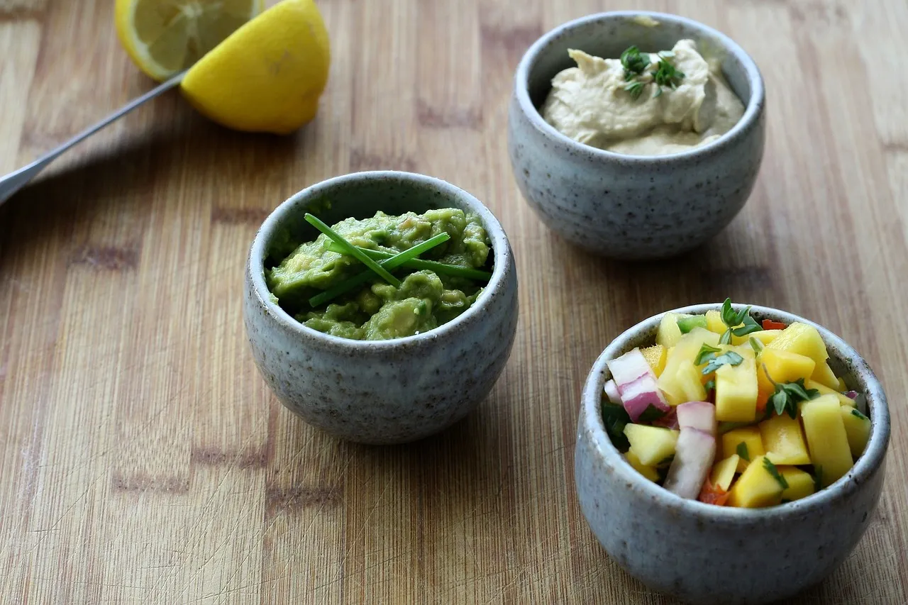 Trzy miseczki z dipami: guacamole, hummus i salsa z mango. Obok połówka cytryny, która może być przyprawą uważaną za afrodyzjak.