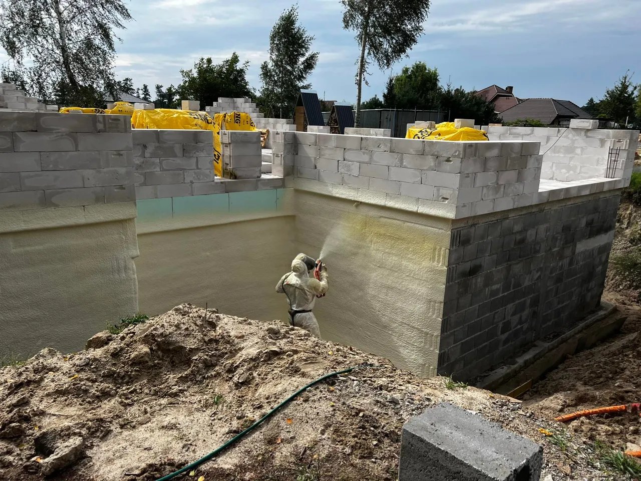 Pracownik w kombinezonie natryskuje piankę, zabezpieczając fundamenty domu. To kluczowy etap budowy.