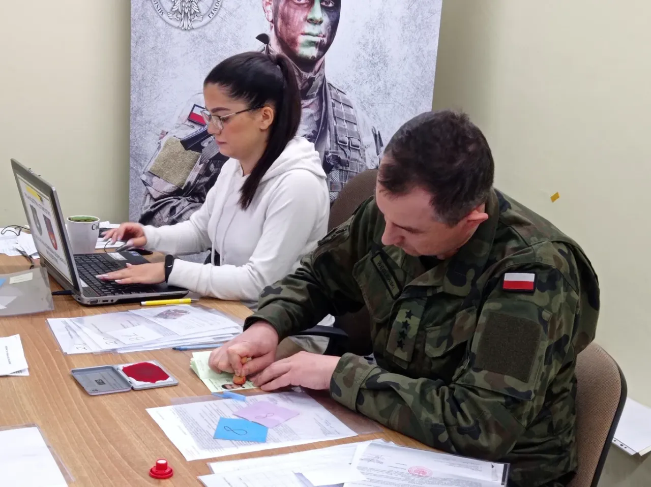 Żołnierz stempluje dokumenty, kobieta pracuje przy laptopie. Tak wygląda badanie na komisji wojskowej.
