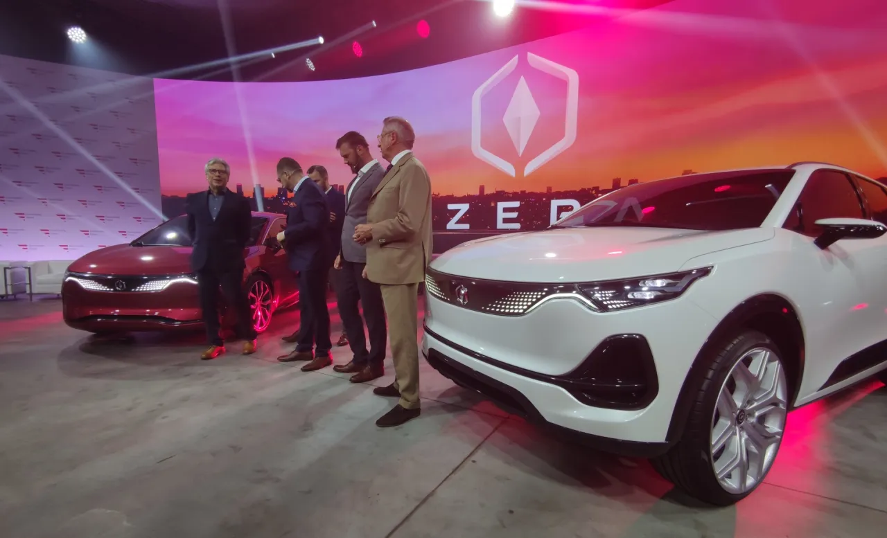 Mężczyźni w garniturach prezentują nowe samochody elektryczne na tle ekranu z logo. To przyszłość polskiego rynku samochodowego.