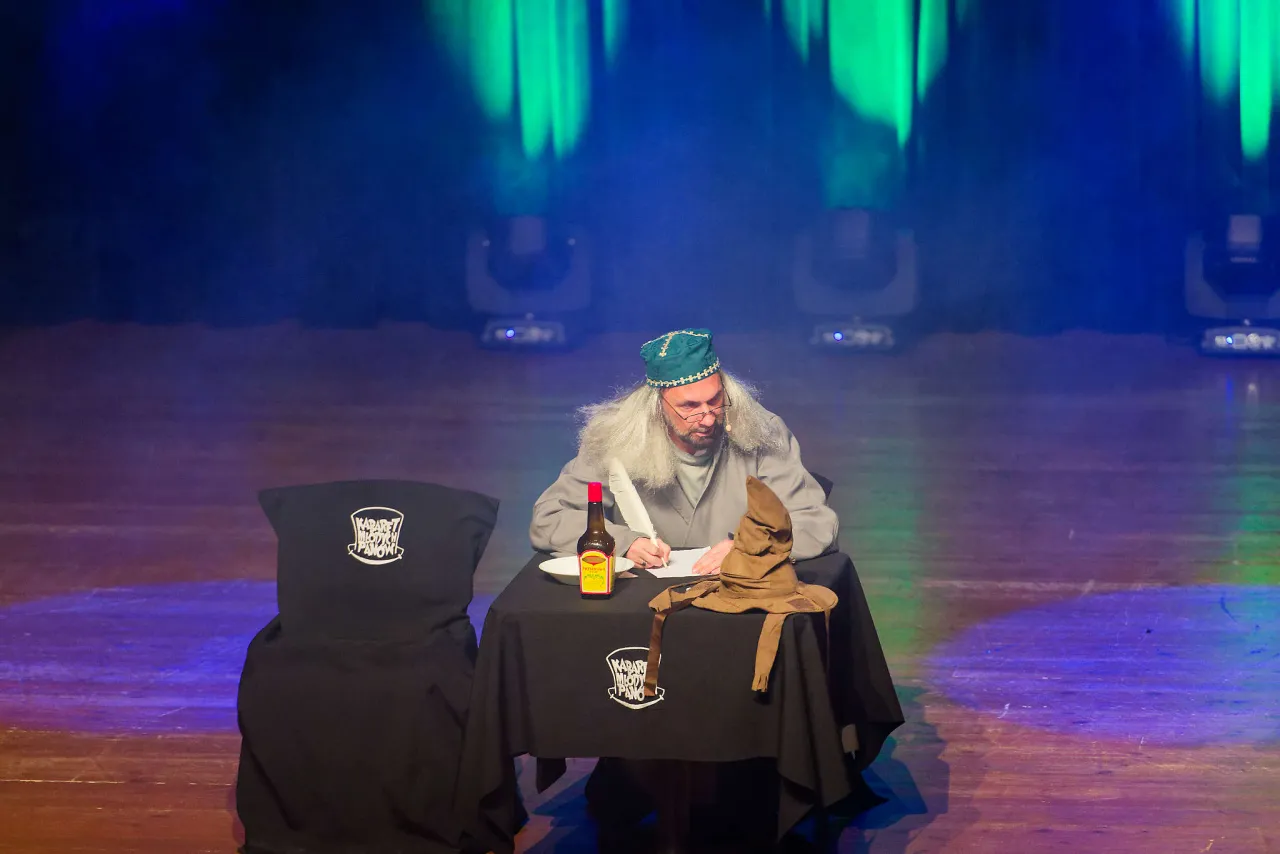 Aktor w peruce i czapce pisze pi&oacute;rem, siedząc przy stole z Tiara Przydziału. Na stole logo Kabaretu Młodych Pan&oacute;w.