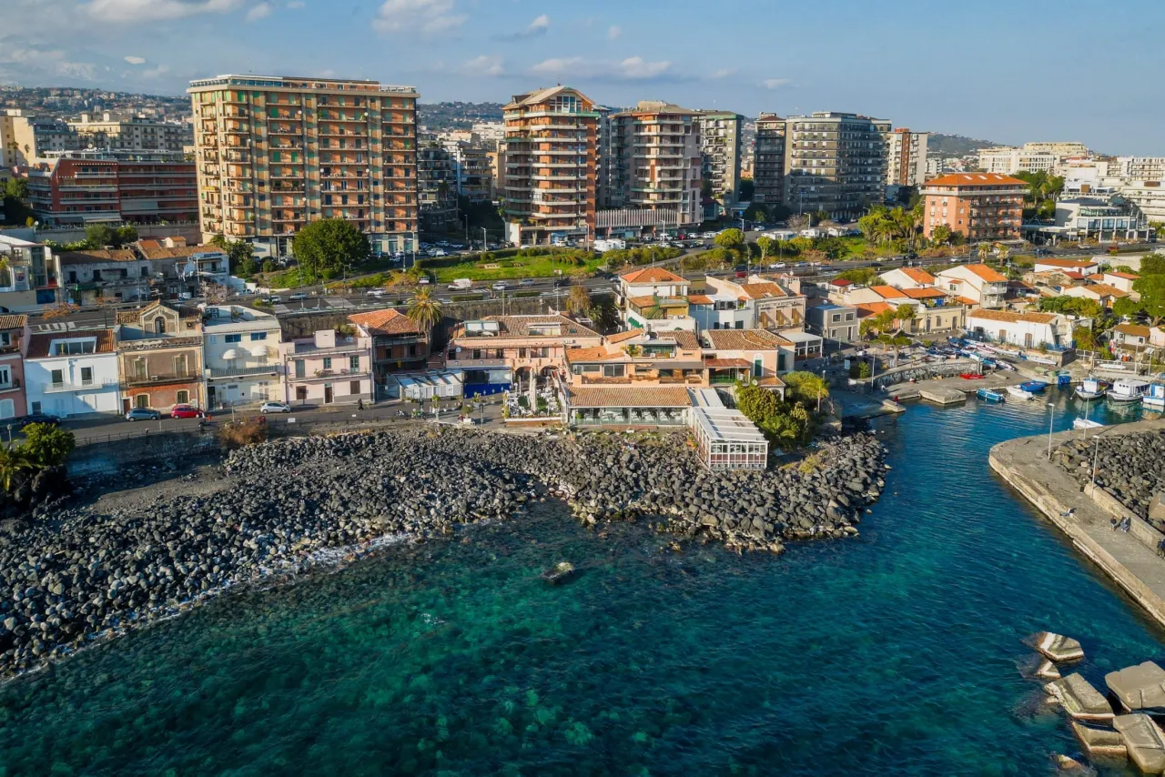 Widok na wybrzeże Katanii z budynkami mieszkalnymi i hotelami. Woda jest turkusowa, a kamieniste wybrzeże tworzy naturalną barierę. Czy w Katanii jest bezpiecznie? Wygląda na spokojne miejsce.