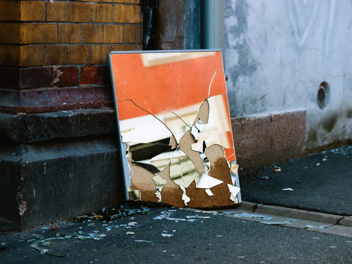 Rozbite lustro leży przy ceglanej ścianie. Zastanawiasz się, gdzie wyrzucić lustro? Uważaj na ostre odłamki szkła.