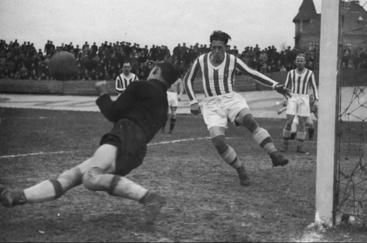 Piłkarz w skoku pr&oacute;buje strzelić gola, bramkarz rzuca się do obrony. To dynamiczne ujęcie pokazuje, skąd pochodzi piłka nożna i jej emocjonującą historię.