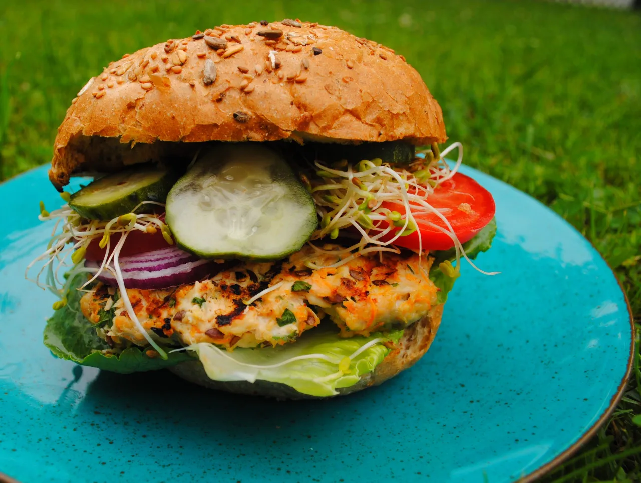Pyszny burger z grilla na talerzu, z bułką pełną ziaren, soczystym kotletem, sałatą, pomidorem, ogórkiem, cebulą i kiełkami.