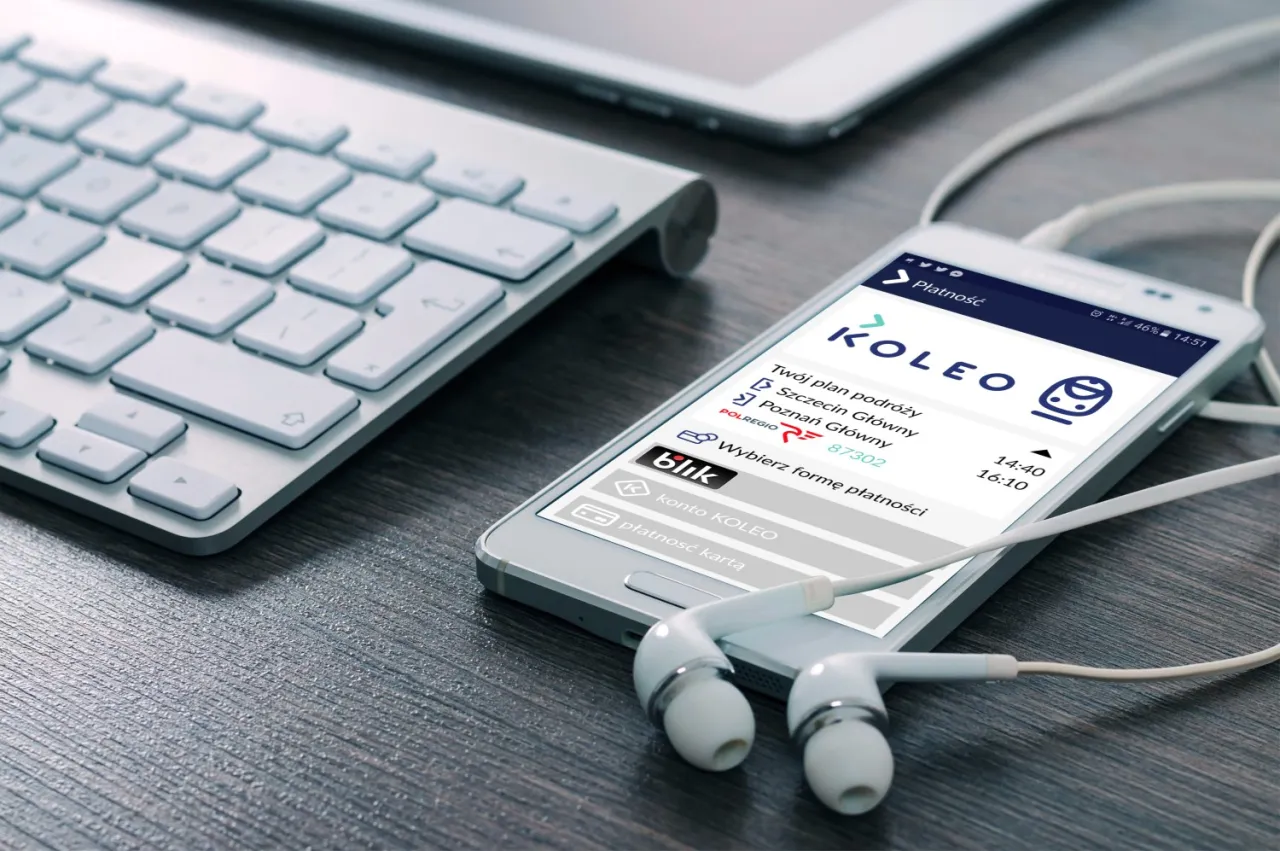 Na ekranie telefonu widać aplikację KOLEO z planem podróży. Co to za pociąg pr? Słuchawki i klawiatura sugerują podróż służbową.