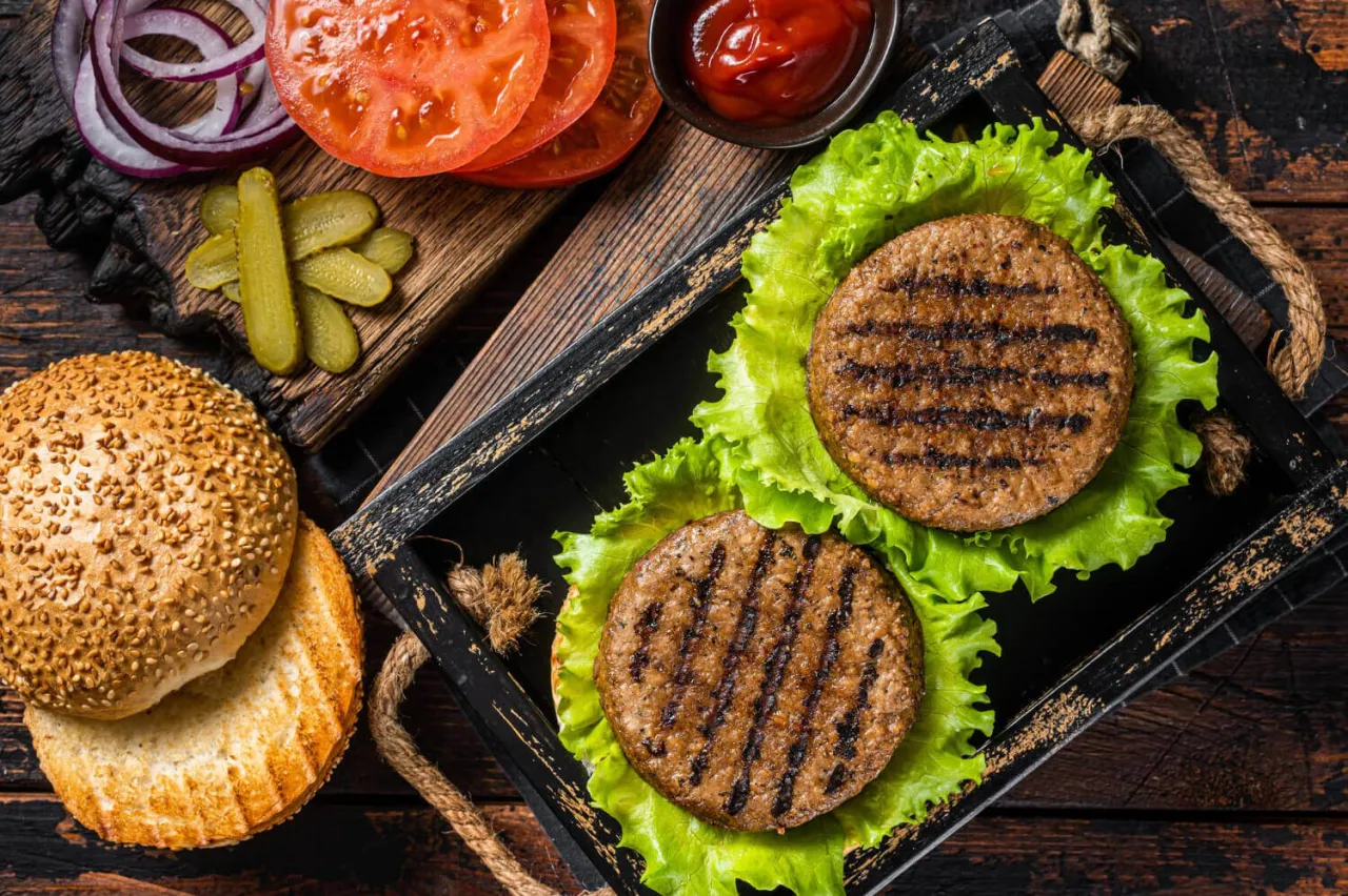 Dwa soczyste kotlety na grilla, ułożone na sałacie, obok bułki, pomidorów, cebuli i pikli. Idealne składniki na pysznego burgera.