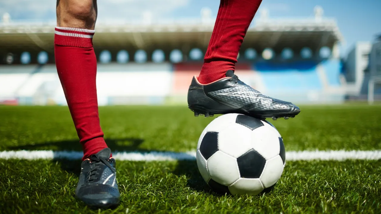 Piłkarz w czerwonych getrach i czarnych butach stoi na boisku, z jedną stopą na piłce. Kto wymyślił piłkę nożną? To pytanie nurtuje wielu fan&oacute;w tej gry.