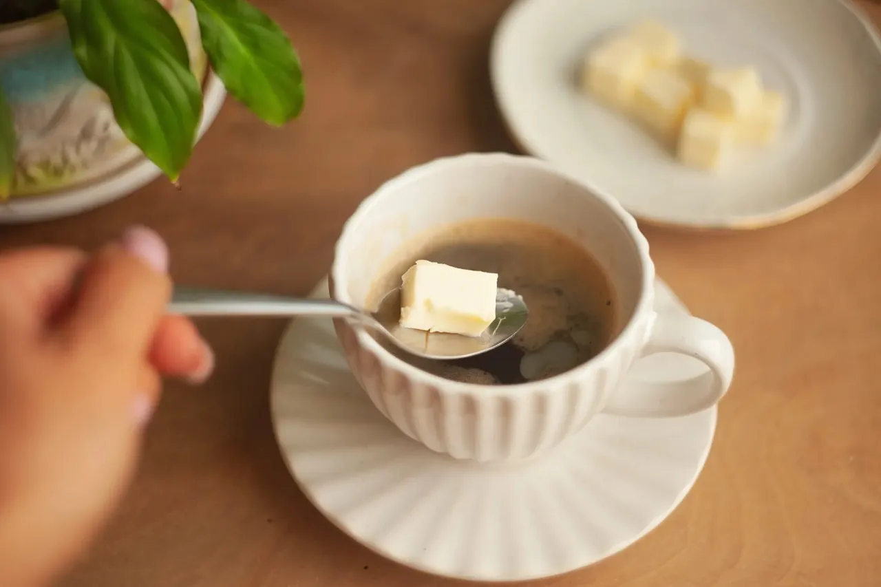 Łyżeczka z kostką masła nad filiżanką kawy. Czy kawa przerywa post przerywany? Na talerzyku obok leżą kolejne kawałki masła.