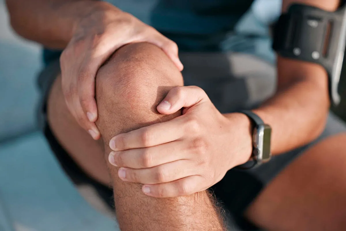 Mężczyzna trzyma się za kolano, co może sugerować rehabilitację po endoprotezie kolana. Na ramieniu ma opaskę sportową, a na nadgarstku zegarek.