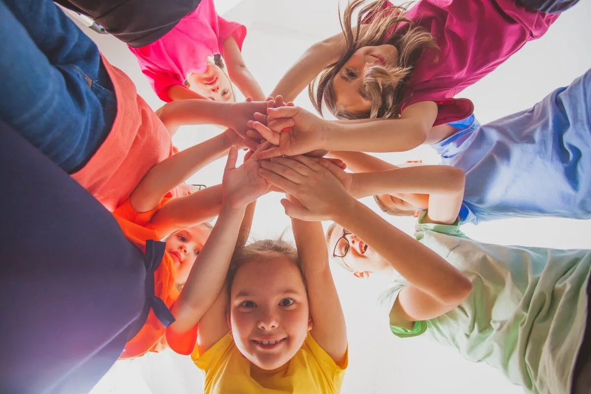 Dzieci i rodzice łączą ręce w geście jedności podczas zajęć integracyjnych w przedszkolu. Radość i współpraca na twarzach.
