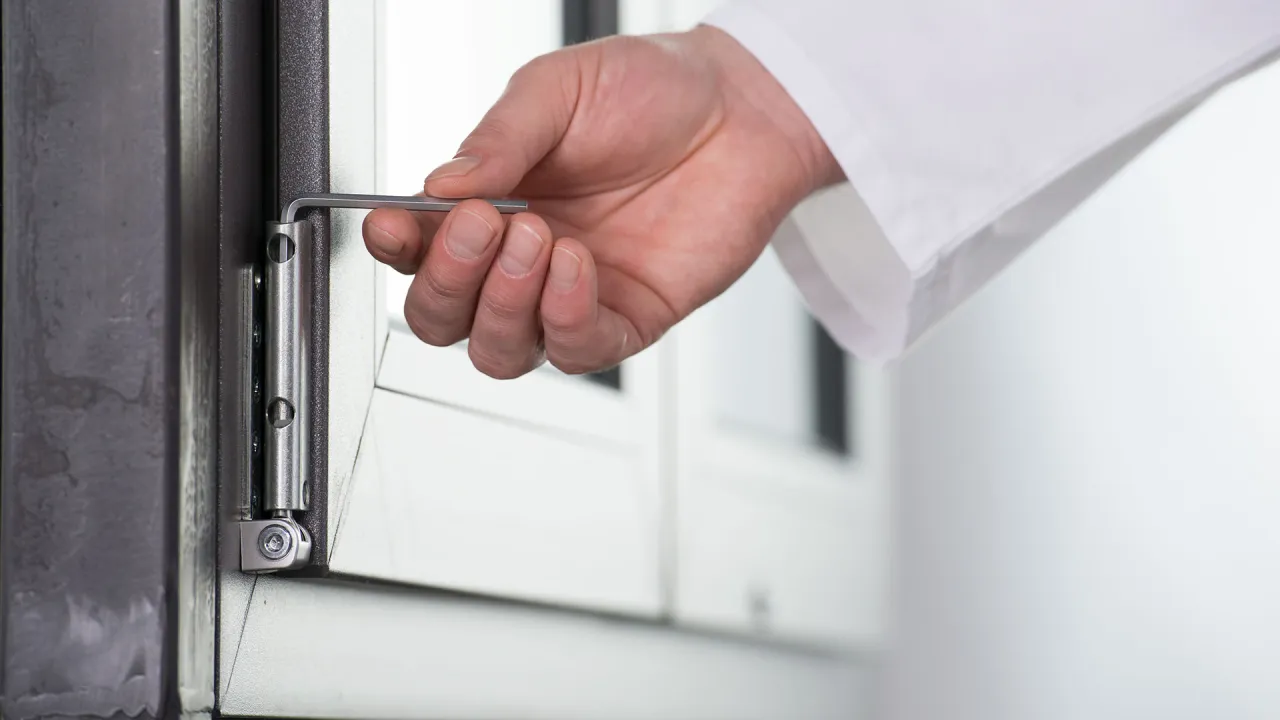 Dłoń z kluczem imbusowym reguluje zawias okna, pokazując, jak zamknąć okno.