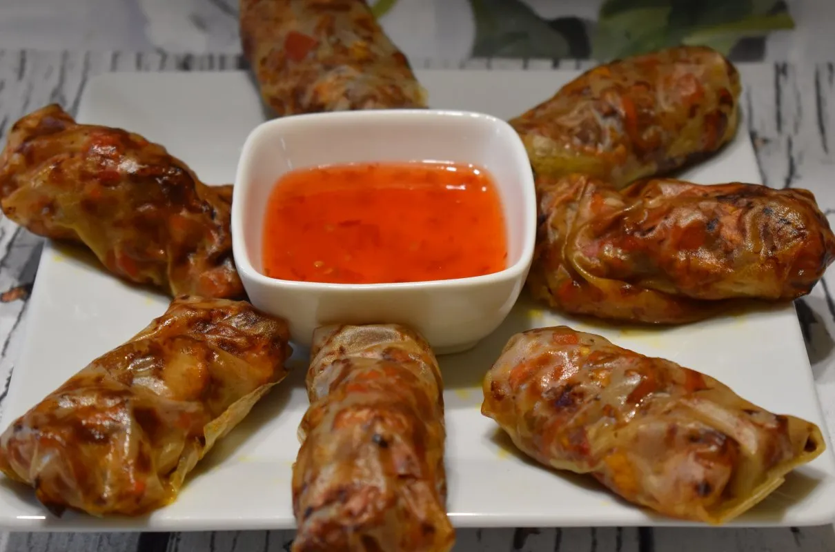 Chrupiące sajgonki z kurczakiem, ułożone wokół miseczki z ostrym, czerwonym sosem. Idealna przekąska.