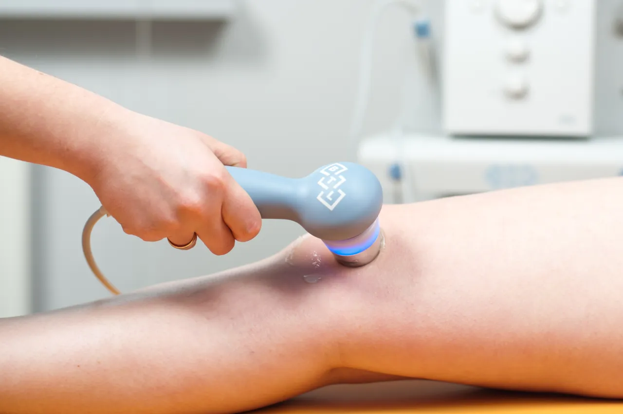 Rehabilitacja ultradźwięki na czym polega? Terapeuta wykonuje zabieg ultradźwiękami na kolanie pacjenta, by przyspieszyć regenerację tkanek.