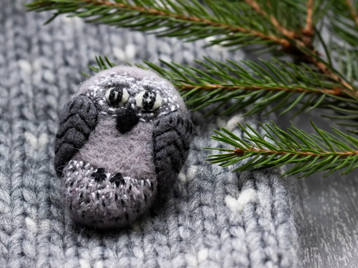 Urocza broszka w kształcie sowy, wykonana z szarej tkaniny z wełny czesankowej, leży na dzianinie. Gałązka świerku dodaje zimowego klimatu.