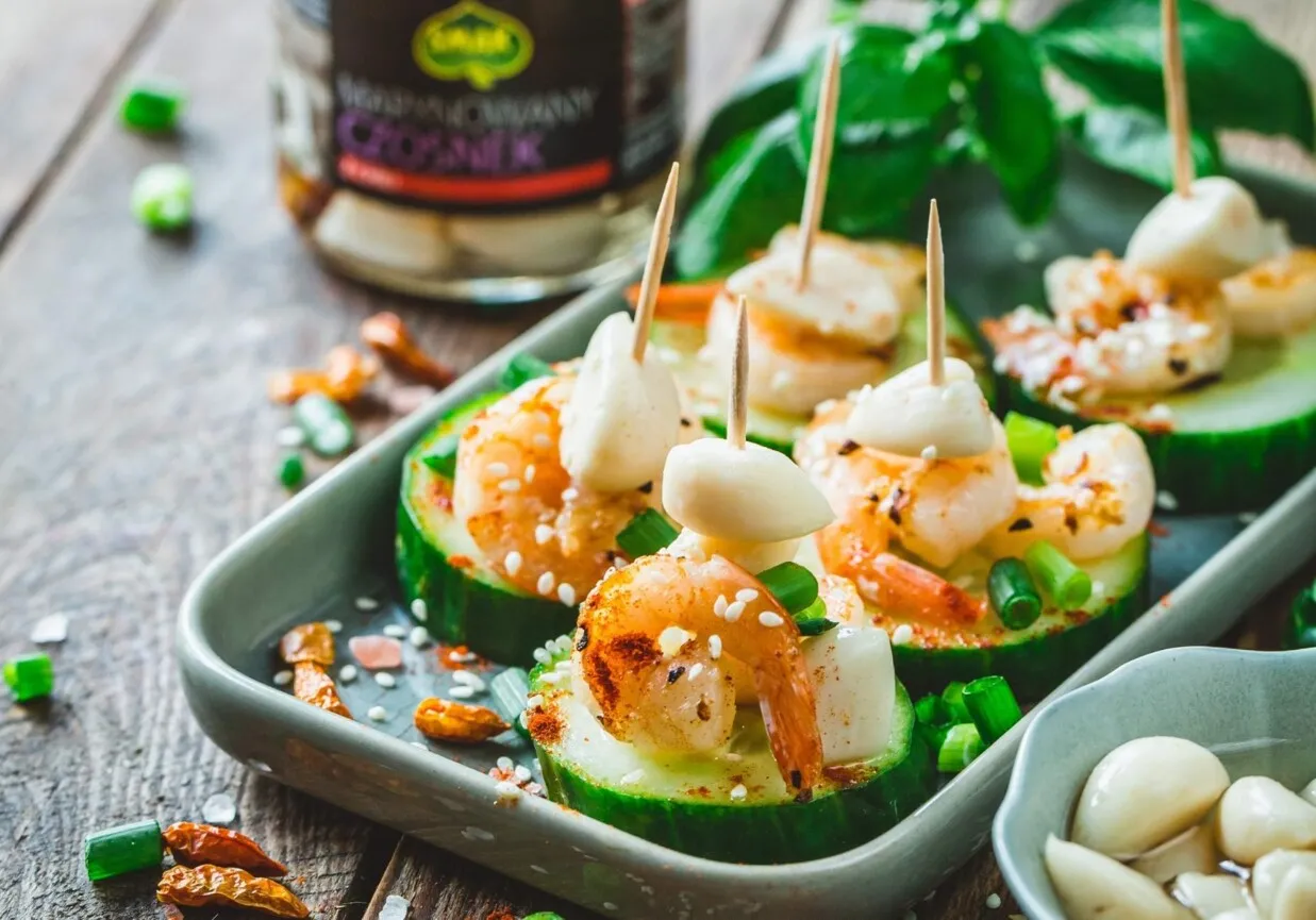 Koreczki z krewetkami na plastrach og&oacute;rka, ozdobione sezamem i szczypiorkiem, z dodatkiem marynowanego czosnku.