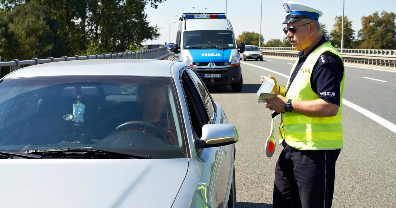 Policjant wypisuje mandat za zbyt wolną jazdę kierowcy srebrnego samochodu na autostradzie. W tle radiow&oacute;z.