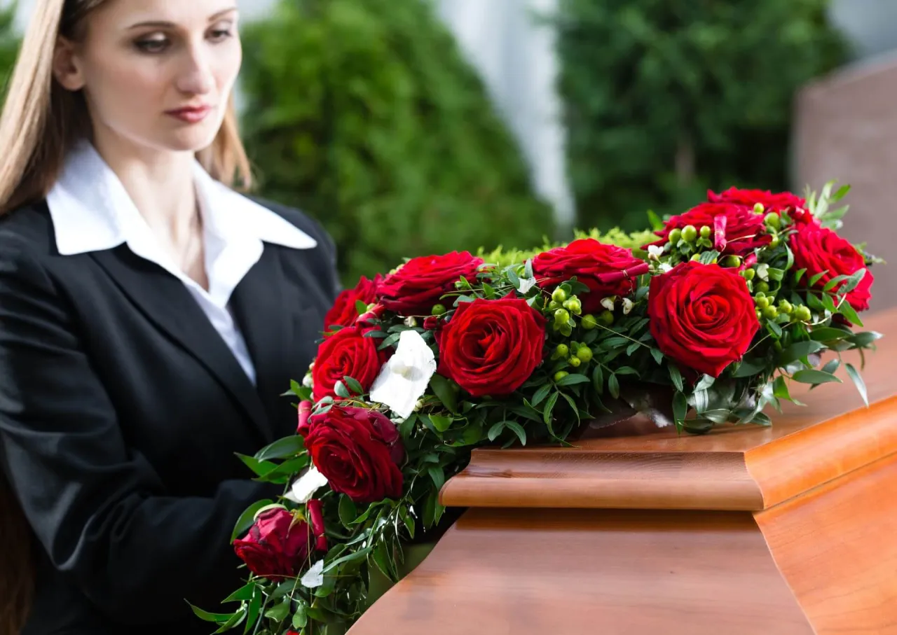 Kobieta w żałobnym stroju kładzie wieniec z czerwonych r&oacute;ż na trumnie. To symboliczne kwiaty na pogrzebie starszej osoby.
