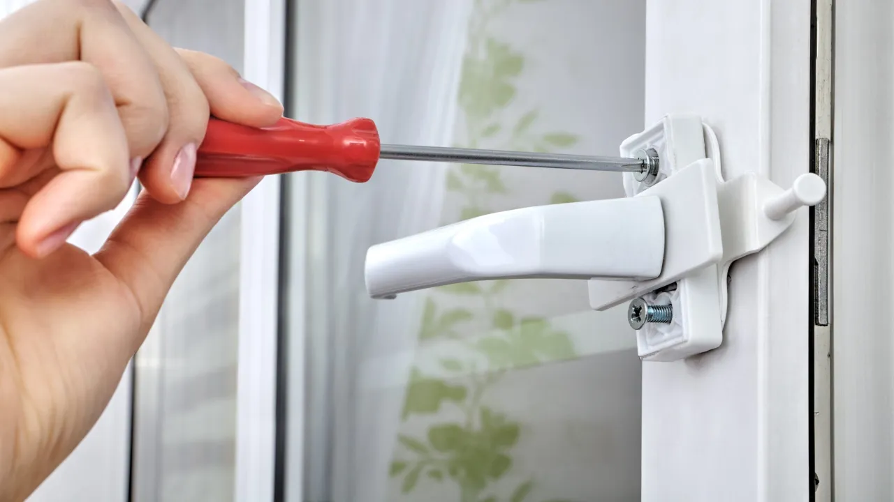 Dłoń z czerwonym śrubokrętem dokręca śrubę w klamce okna, pokazując, jak zamknąć okno.