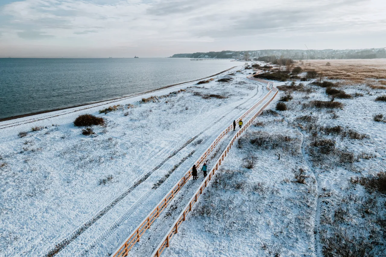Spacer po zaśnieżonych ścieżkach w rezerwacie przyrody Mechelińskie Łąki. Widać drewnianą kładkę, morze i kilka os&oacute;b spacerujących.