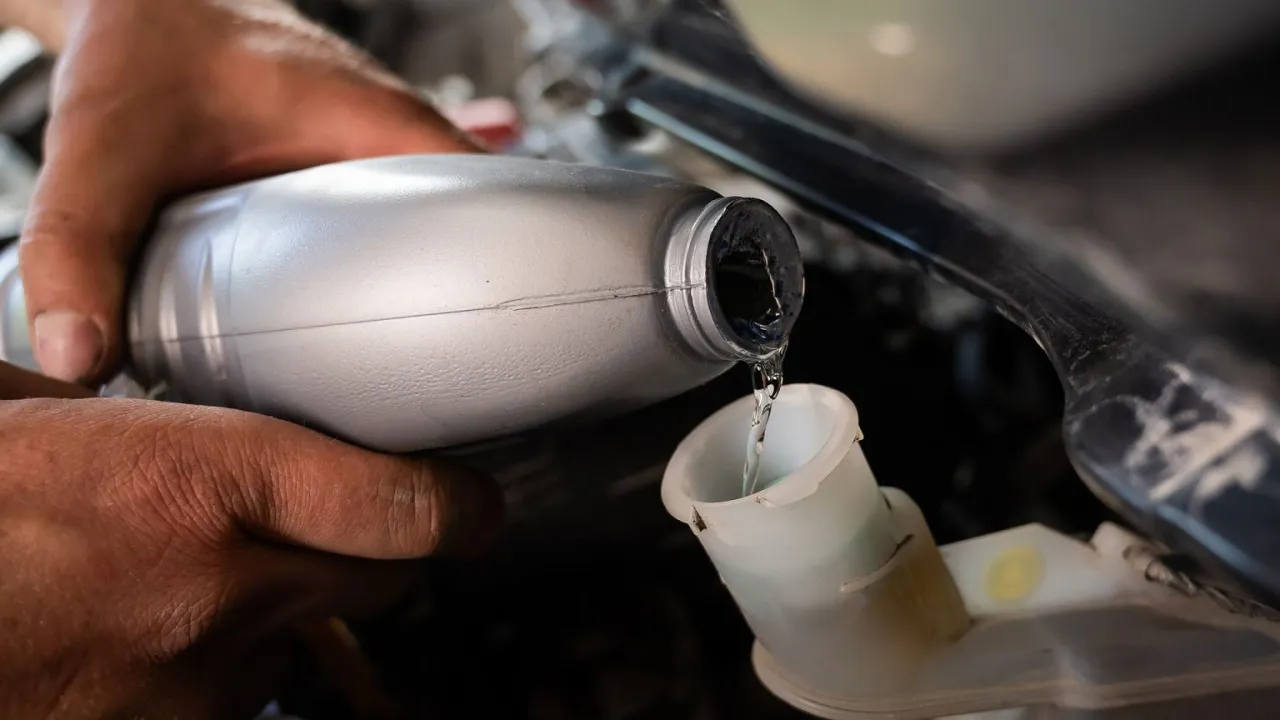 Dłoń przelewa płyn ze srebrnej butelki do zbiorniczka. Pamiętaj, jak często wymieniać olej w samochodzie, by silnik działał sprawnie.