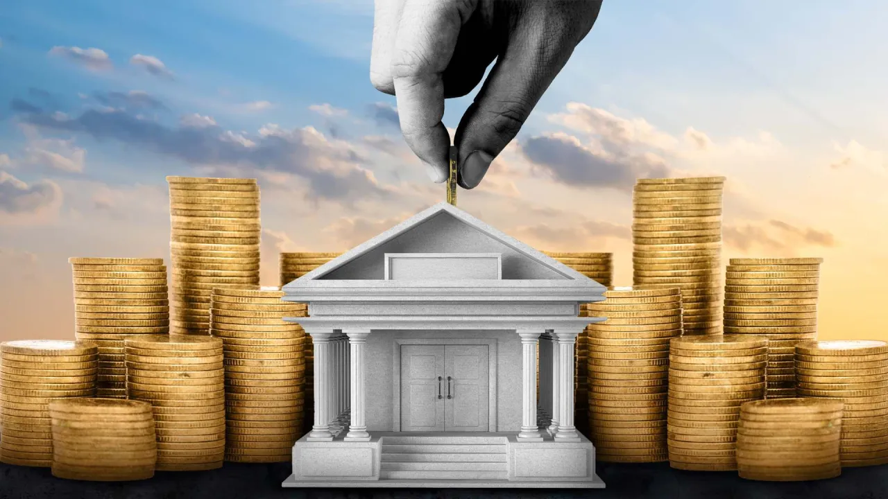 Dłoń wrzuca monetę do banku, otoczonego stosami złota. Symbolizuje to bezpieczeństwo depozytów, o które pyta bankowy fundusz gwarancyjny jakie banki.