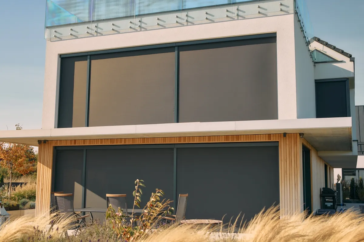 Nowoczesny dom z dużymi oknami, chronionymi przez eleganckie rolety fasadowe zewnętrzne. Na tarasie stolik i krzesła.