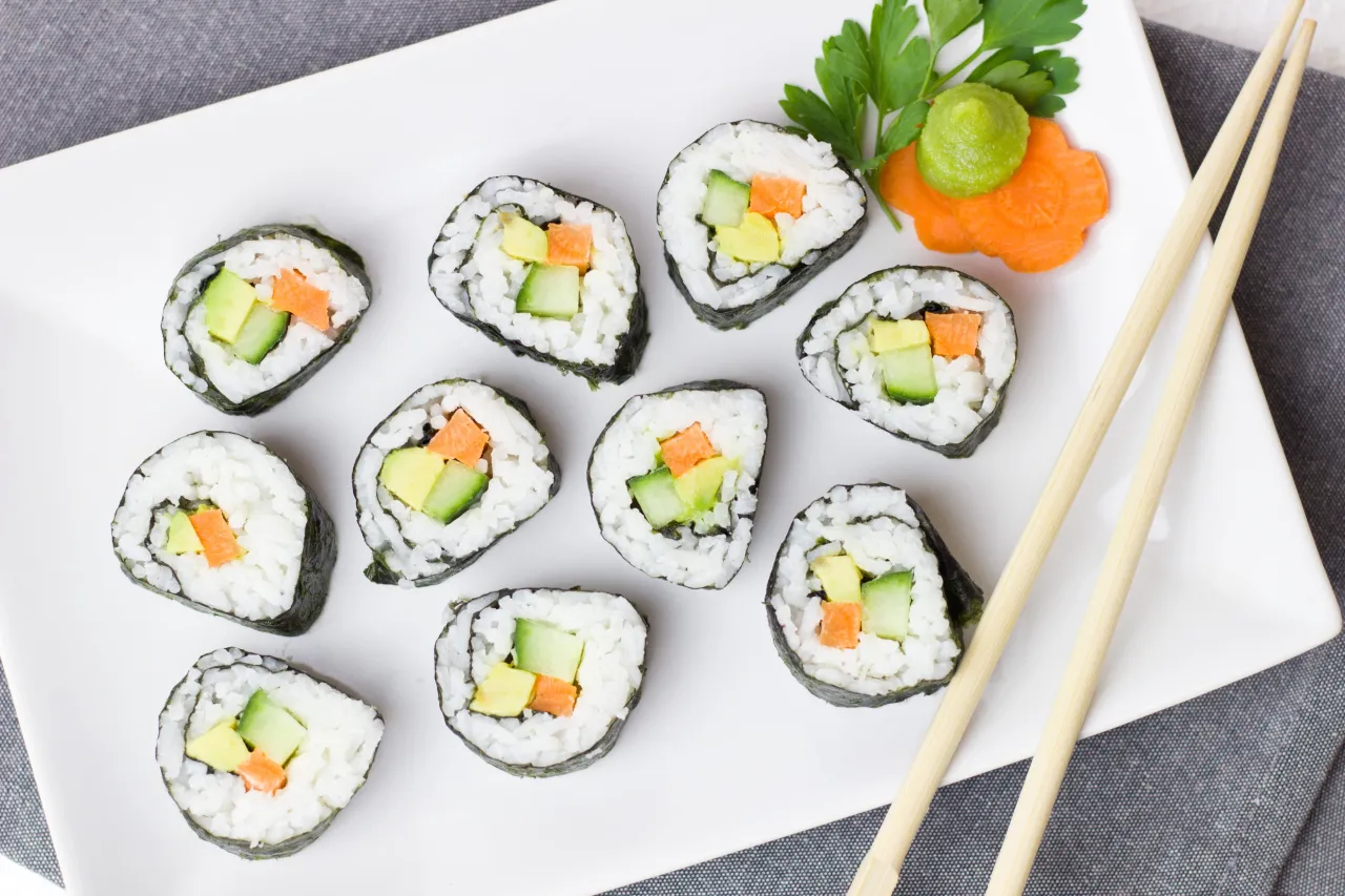 Roladki sushi z awokado, og&oacute;rkiem i marchewką. Obok wasabi i ozdobna marchewka. Pałeczki czekają na rybę do sushi.