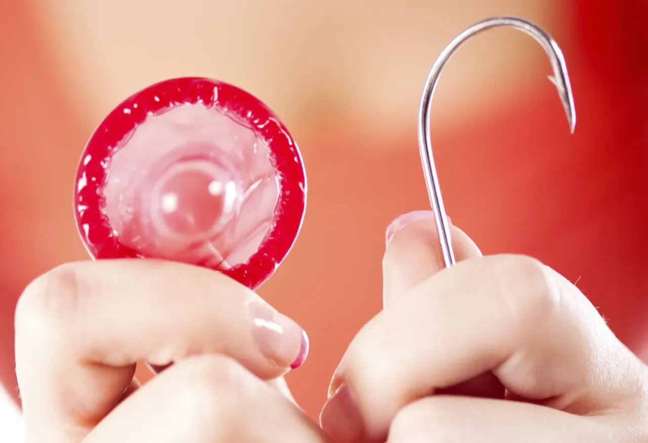 Dłonie trzymają prezerwatywę i haczyk. Czy wytrysk w prezerwatywie jest bezpieczny? Haczyk symbolizuje ryzyko.