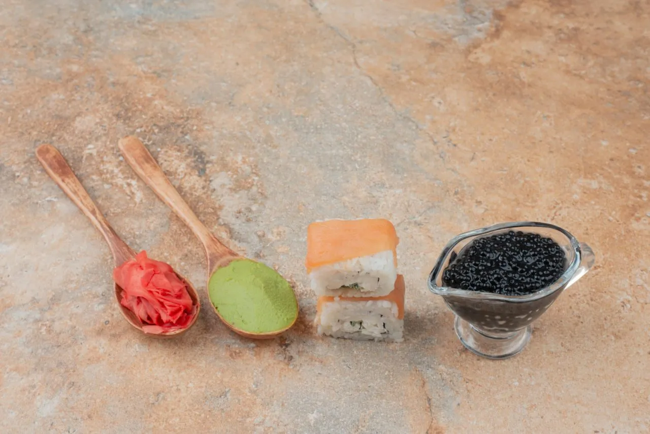 Dwa kawałki sushi z łososiem, obok łyżki z zielonym wasabi, łyżki z marynowanym imbirem i sosjerki z czarnym kawiorem.