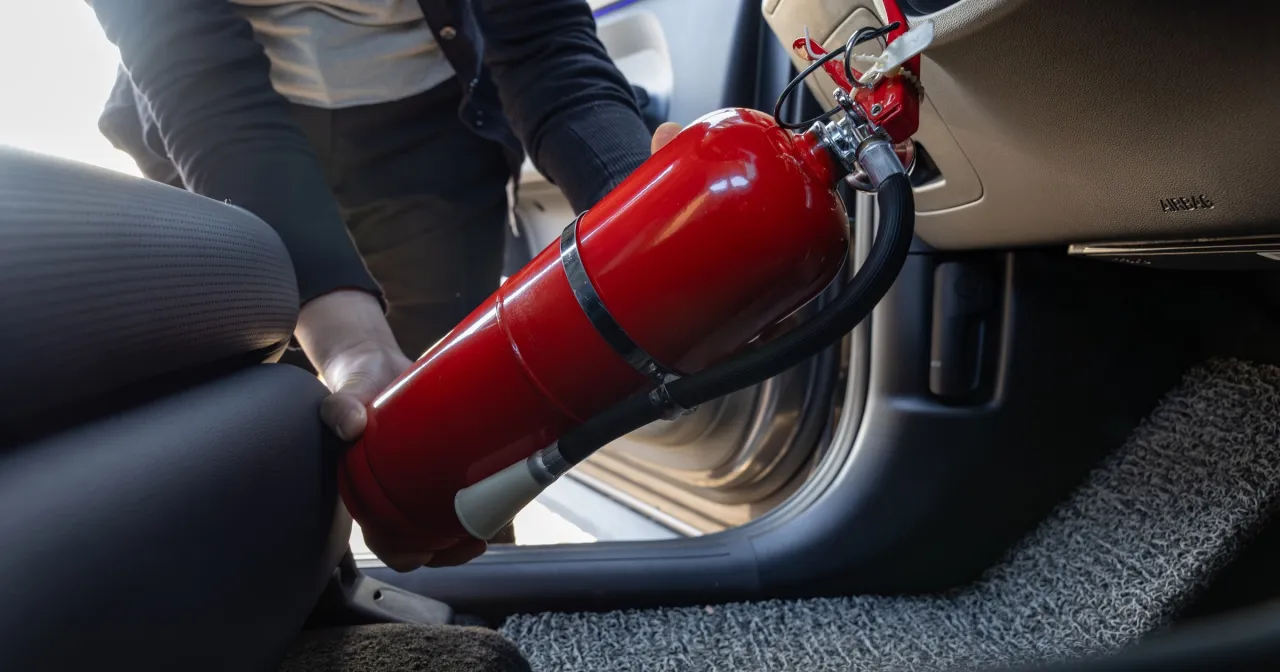 Ręce wyjmujące czerwoną gaśnicę z samochodu. Brak gaśnicy może skutkować mandatem.
