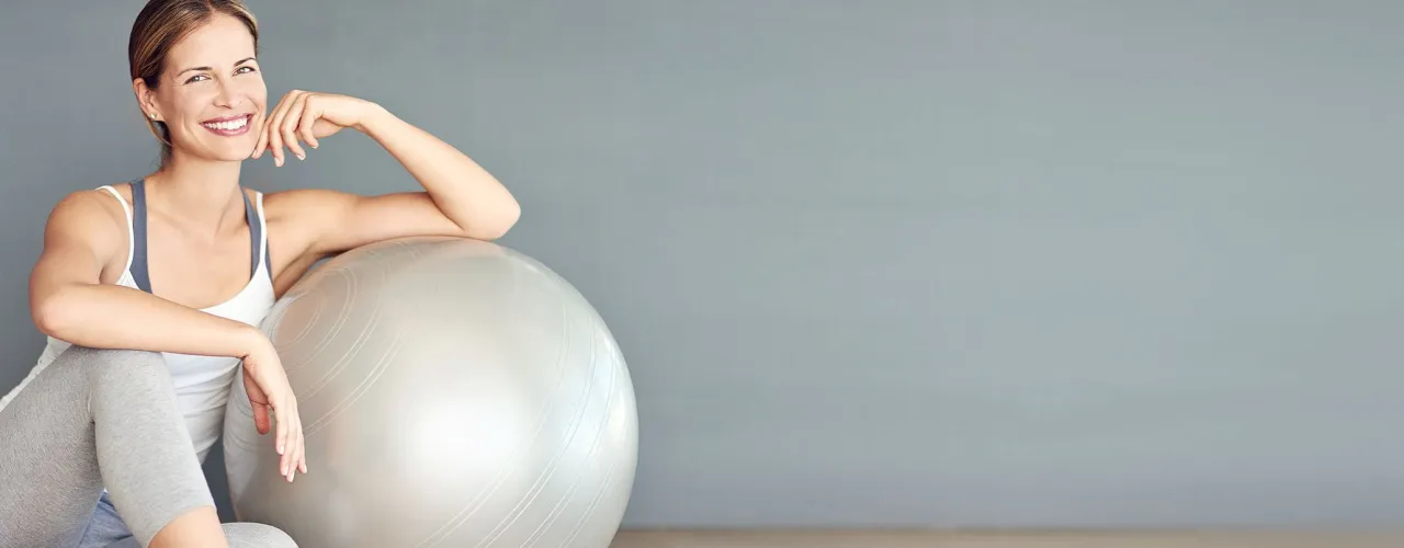 Uśmiechnięta kobieta w stroju sportowym opiera się o dużą, szarą piłkę gimnastyczną. Dowiedz się, jak ćwiczyć na piłce żeby schudnąć i poczuć się lepiej!