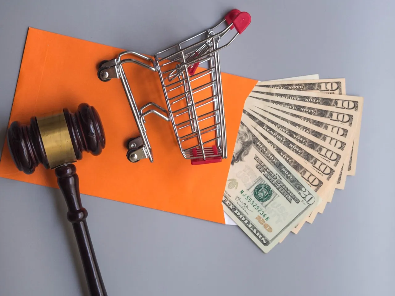 Młotek sędziowski, koszyk sklepowy i banknoty na pomarańczowej kopercie. Czy warto ogłosić upadłość konsumencką?