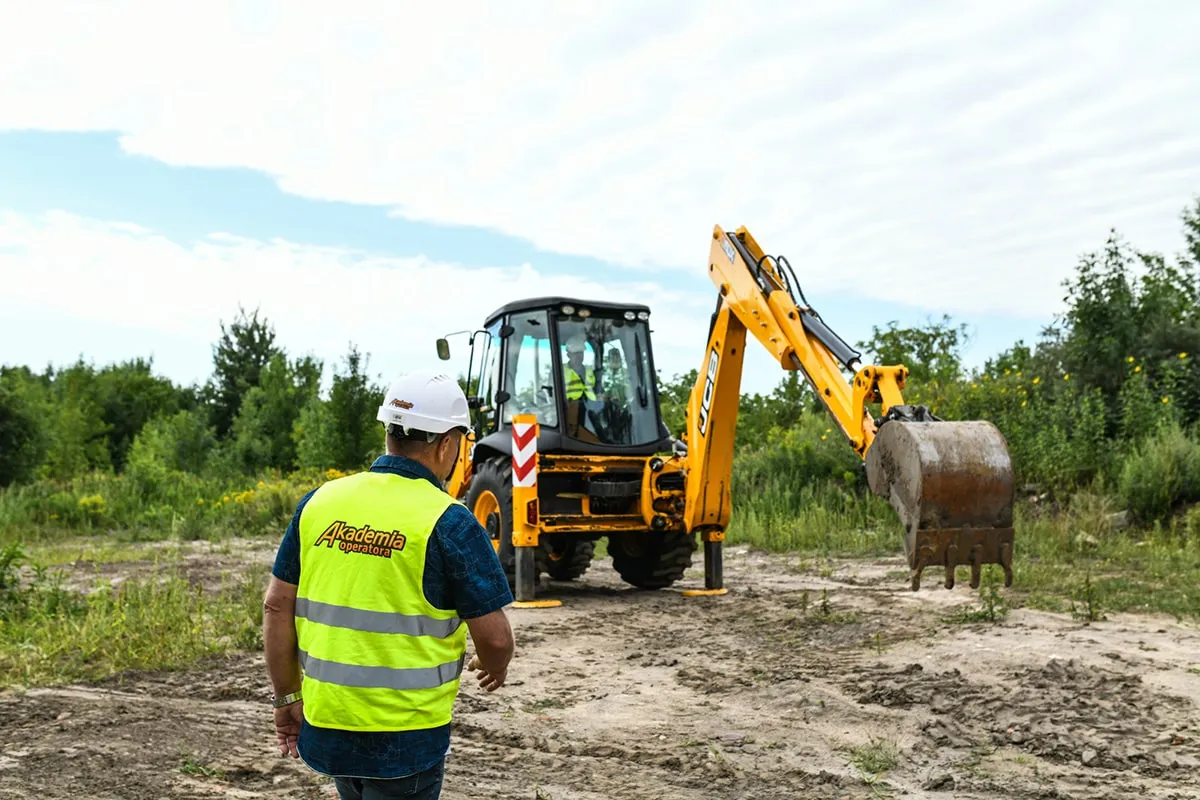 Operator w kamizelce odblaskowej i kasku, przed koparko-ładowarką. Przygotowanie do egzaminu na ładowarkę, pytania i odpowiedzi.