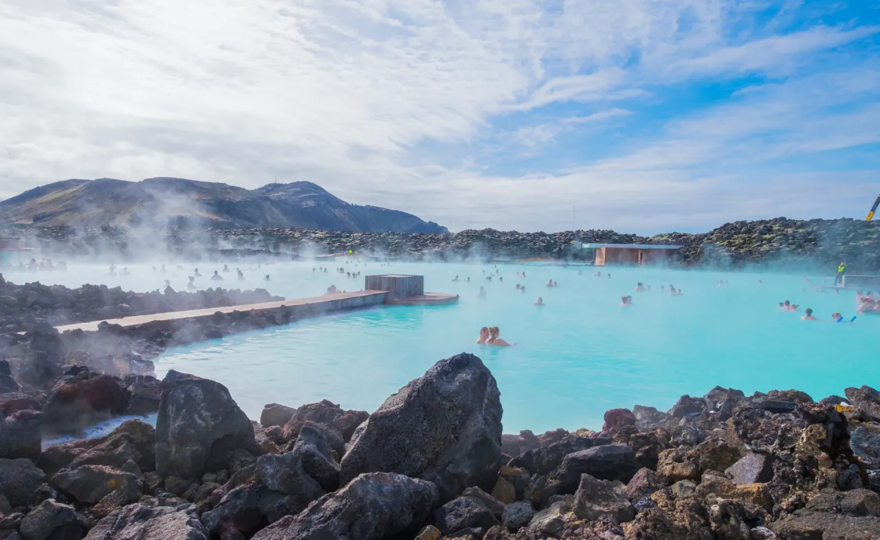 Para ludzi relaksujących się w turkusowej wodzie geotermalnego basenu Blue Lagoon, otoczonego wulkanicznymi skałami i parą.