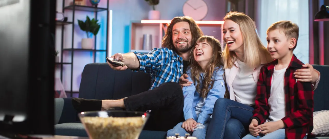Rodzina śmieje się, oglądając telewizję. Dofinansowanie do dekodera dla kogo? Dla wszystkich, kt&oacute;rzy chcą cieszyć się wsp&oacute;lnymi chwilami.