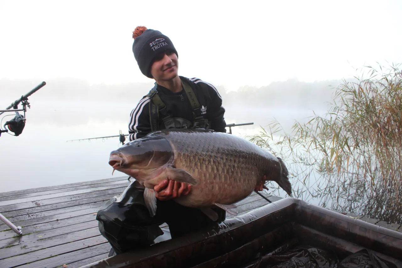 Młody wędkarz z okazałym karpiem na rękach. W tle mglisty poranek nad jeziorem, wędki i trzciny. Idealna jaka długość wędki na spinning na takie połowy?