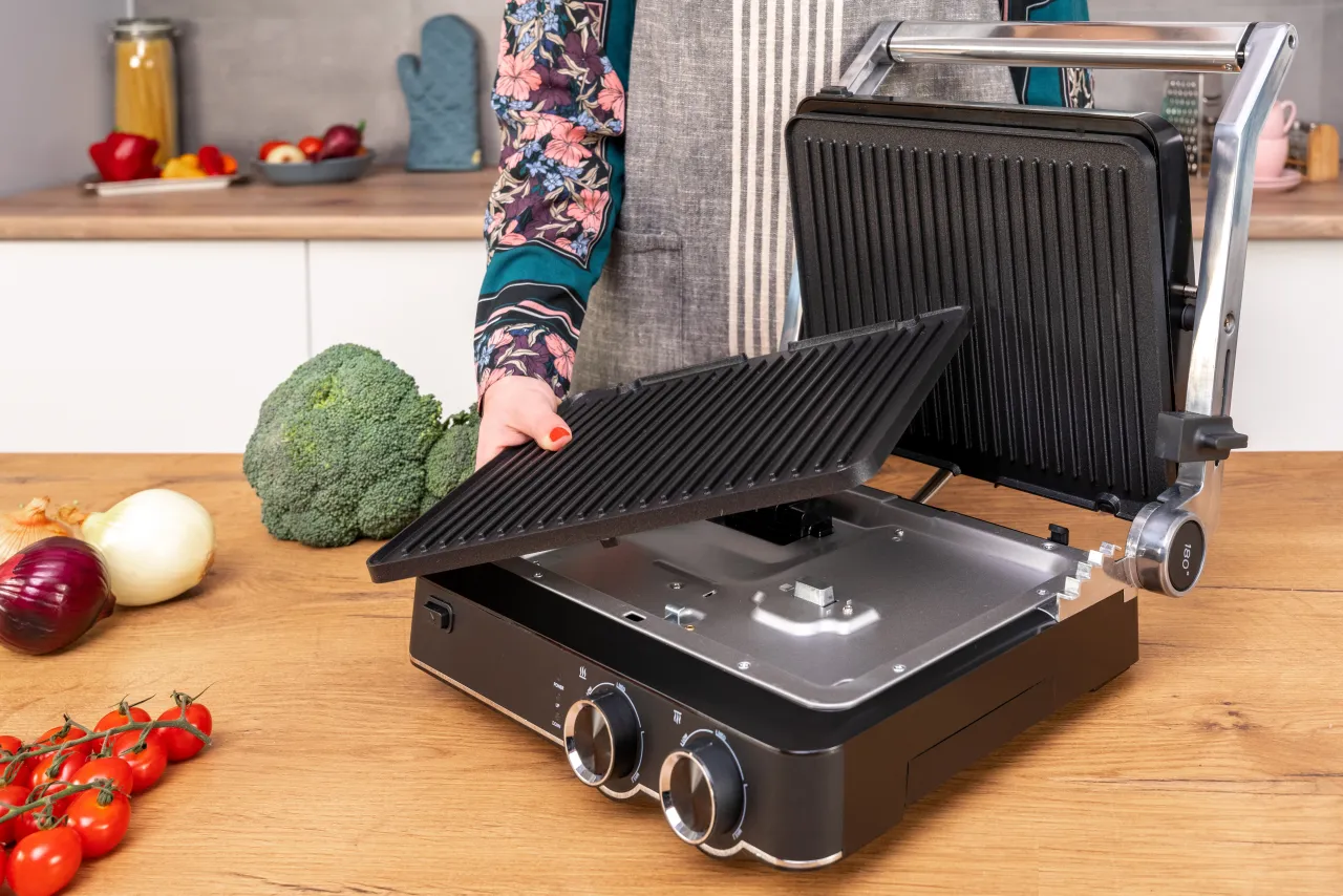 Osoba otwiera grill elektryczny Götze & Jensen GL802X, obok leżą warzywa: brokuł, pomidorki, cebule.