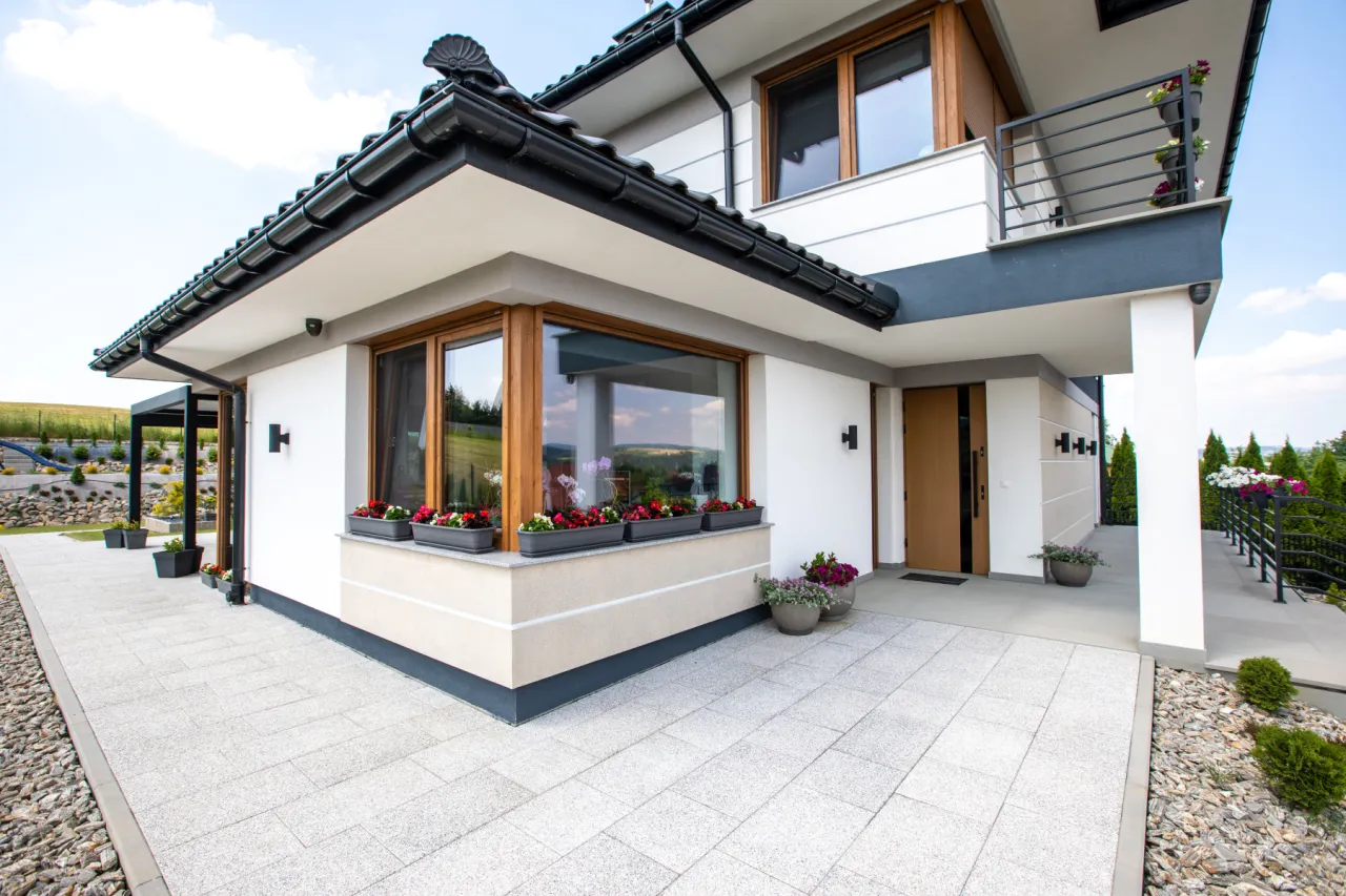 Nowoczesny dom z dużym oknem narożnym, ozdobionym kwiatami. Obok wejścia stoi betonowy słup, a wokół domu piękne patio.