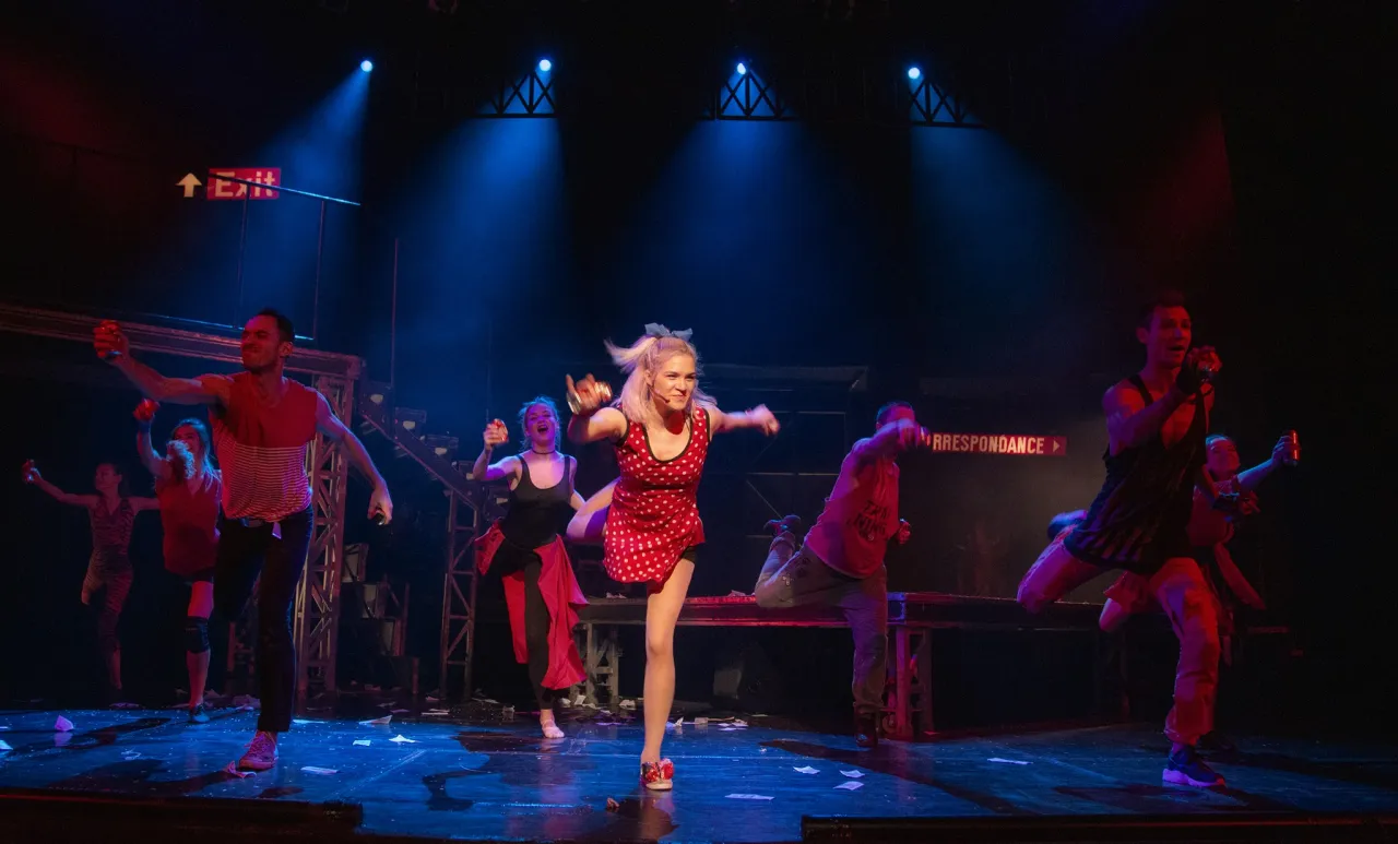 Tancerze na scenie, w tym kobieta w czerwonej sukience w kropki, żywiołowo tańczą. Czy to scena z musicalu 