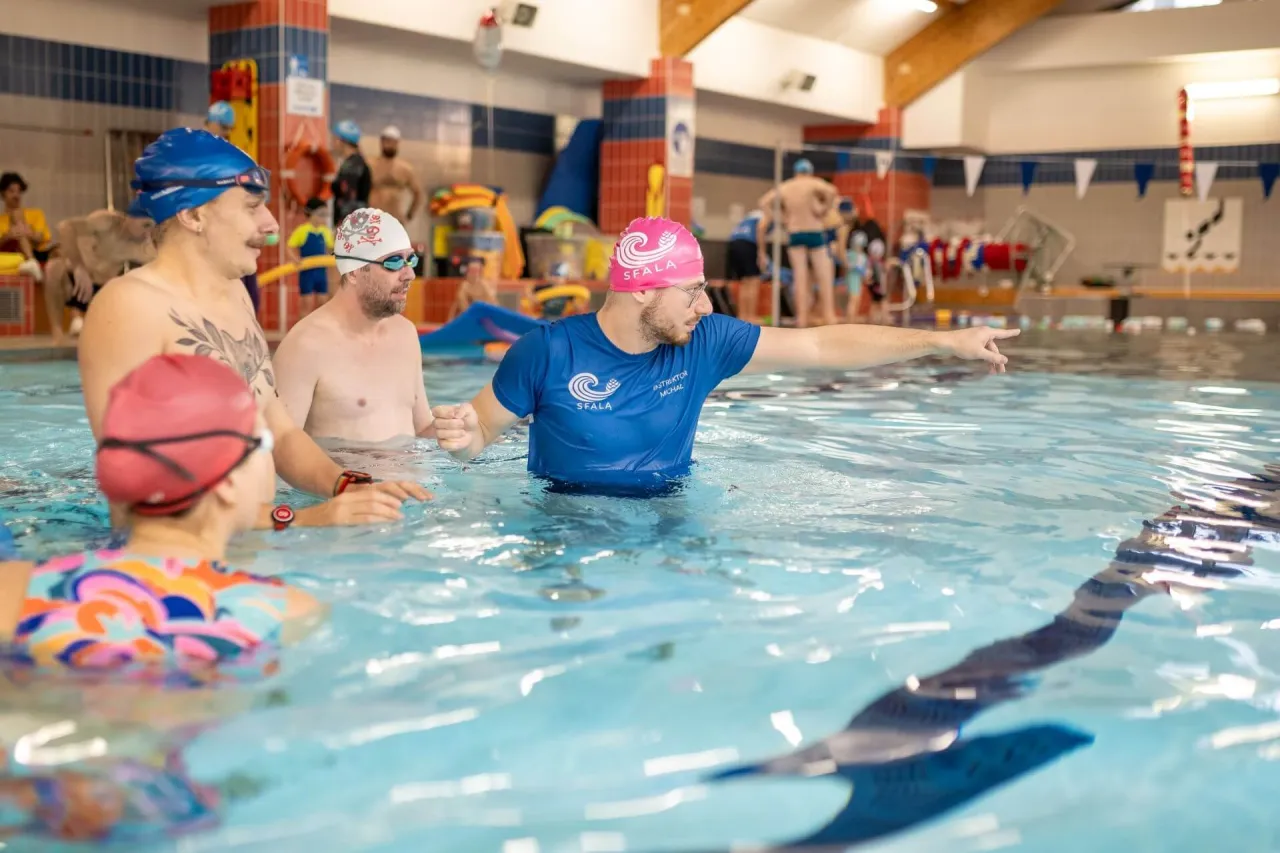 Instruktor pokazuje, jak nauczyć pływać dorosłego. Grupa osób w basenie, w tym mężczyźni i kobieta, słucha wskazówek.