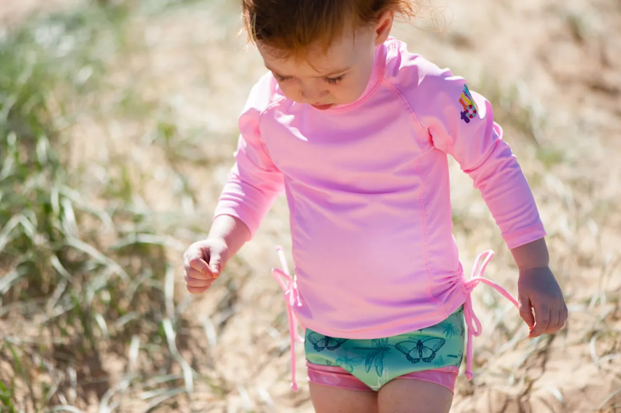 Mała dziewczynka w r&oacute;żowej bluzce do pływania z motylkami na spodenkach bawi się na plaży.
