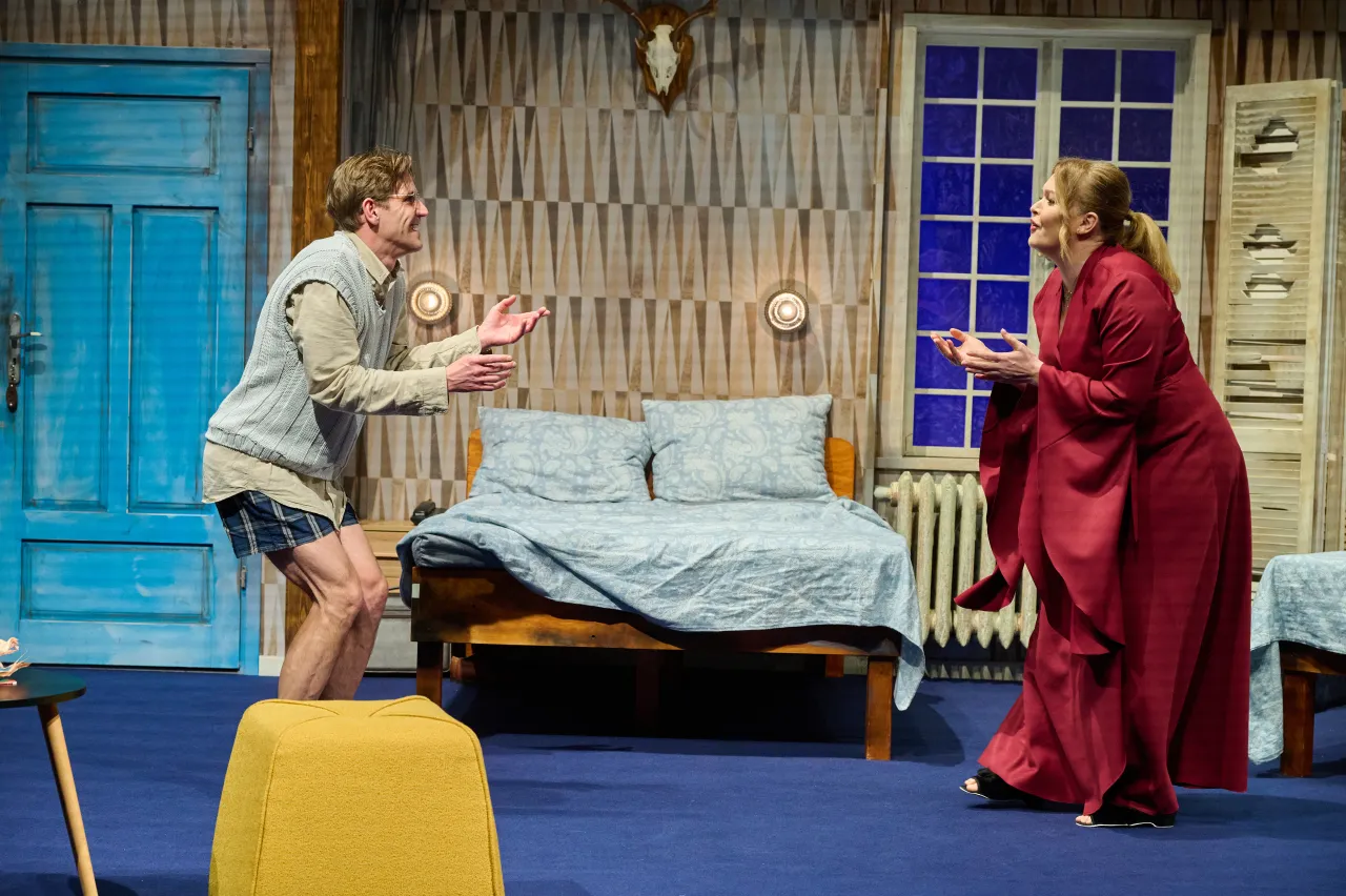 Aktorzy na scenie, mężczyzna w szortach i kobieta w czerwonej sukni, gestykulują przy łóżku pełnym cudzoziemców.