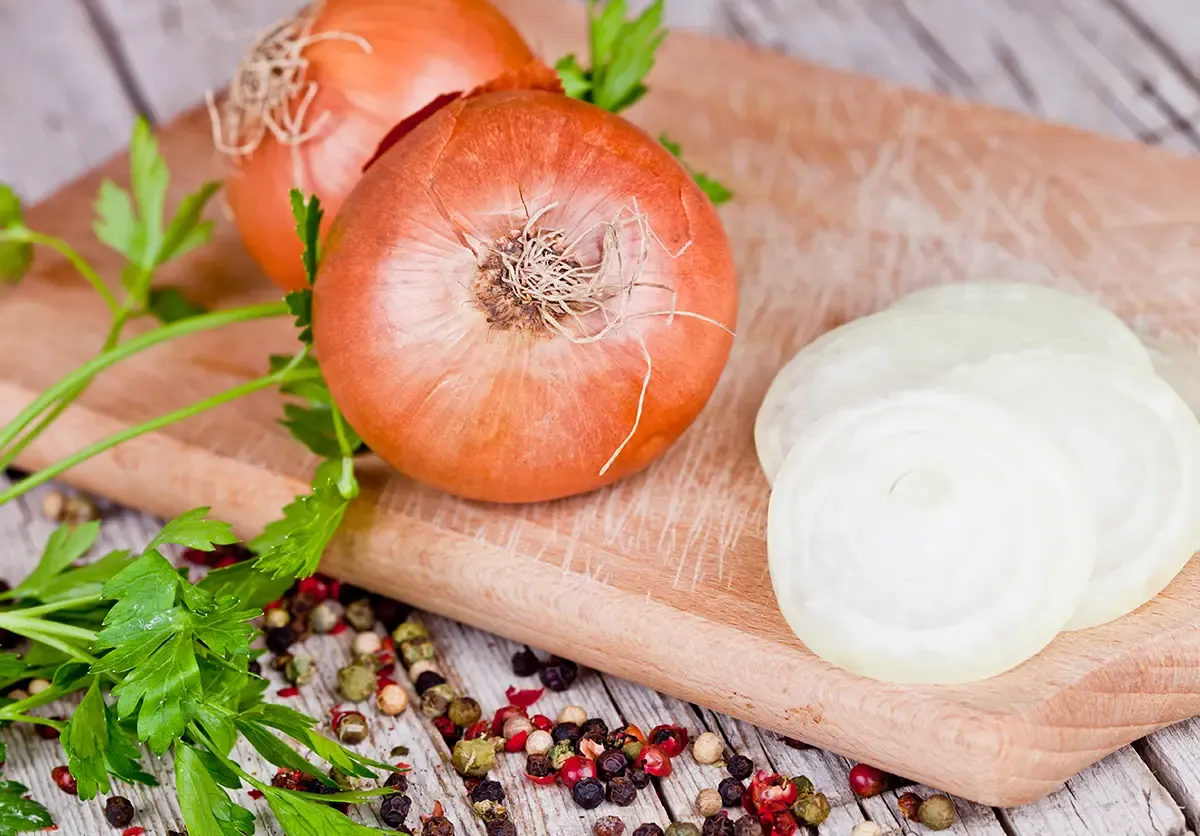 Dwie całe cebule i kilka plasterków na desce, obok natka pietruszki i kolorowy pieprz. Czy można mrozić cebulę? Tak, można ją mrozić.